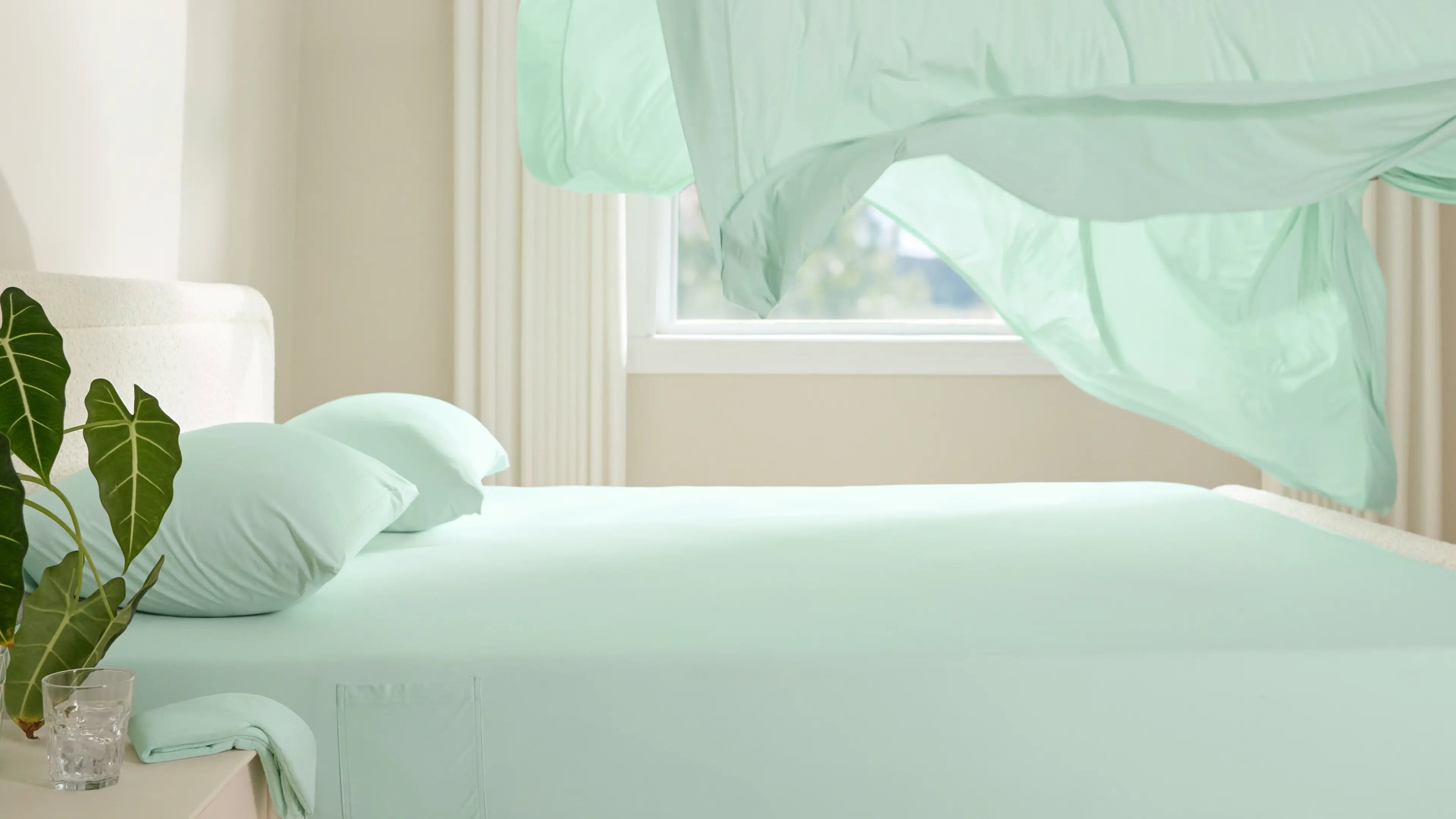 A light green cooling sheet billows over a bed in a bright, airy room, showcasing its lightweight and breathable fabric