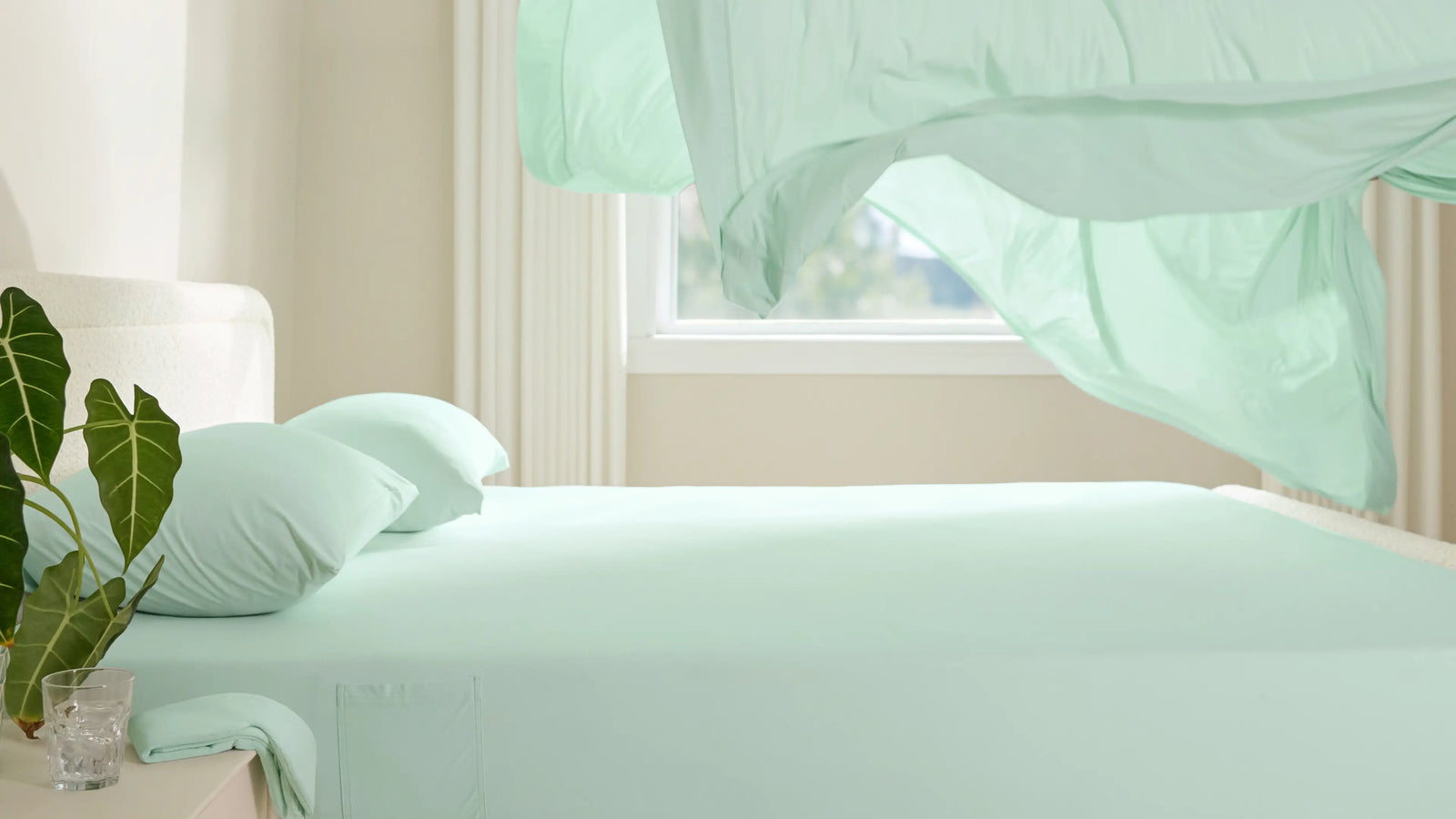 A light green cooling sheet billows over a bed in a bright, airy room, showcasing its lightweight and breathable fabric
