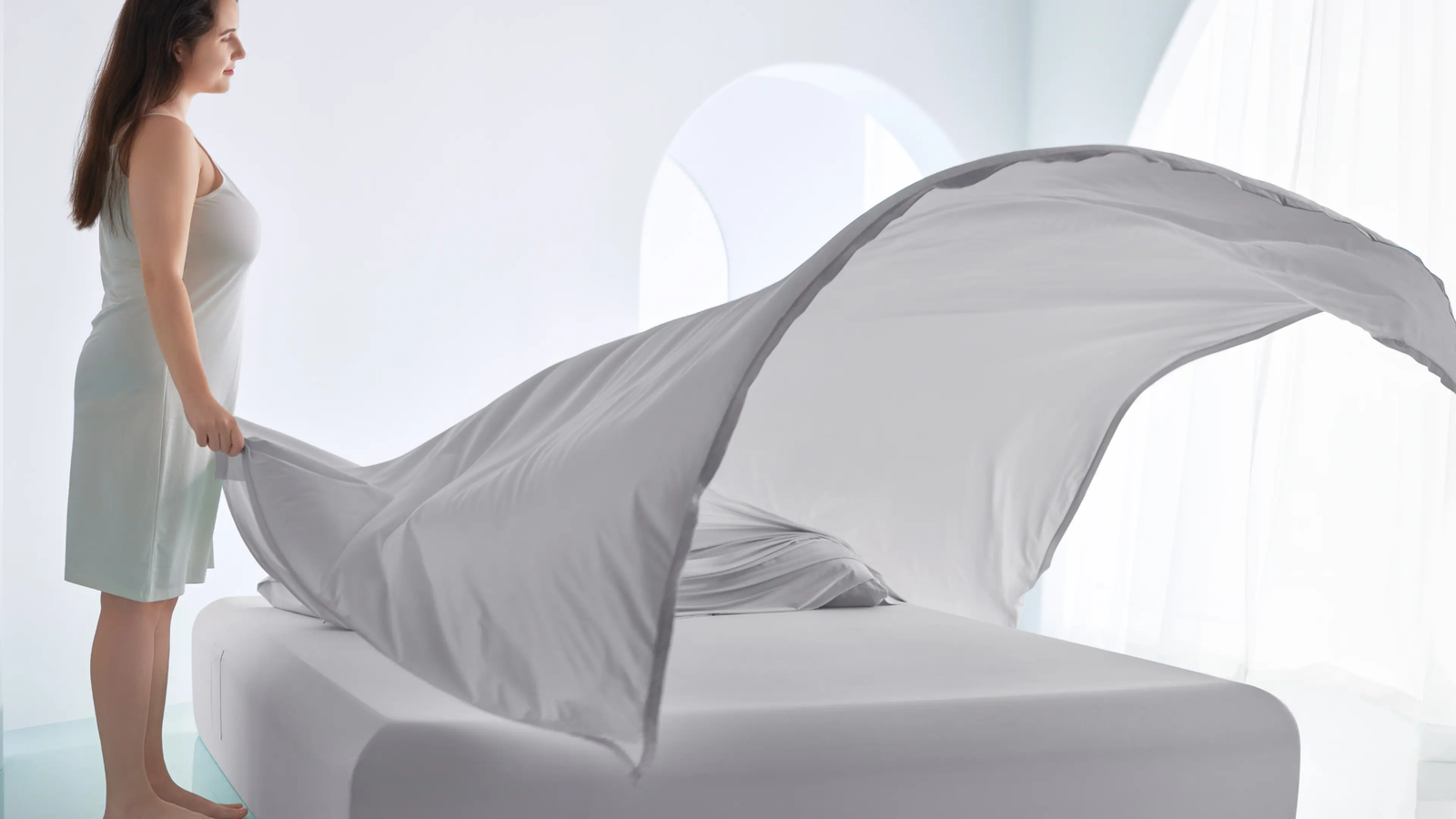 A woman in a nightgown spreads a large, billowing white sheet over a bed in a bright, airy bedroom