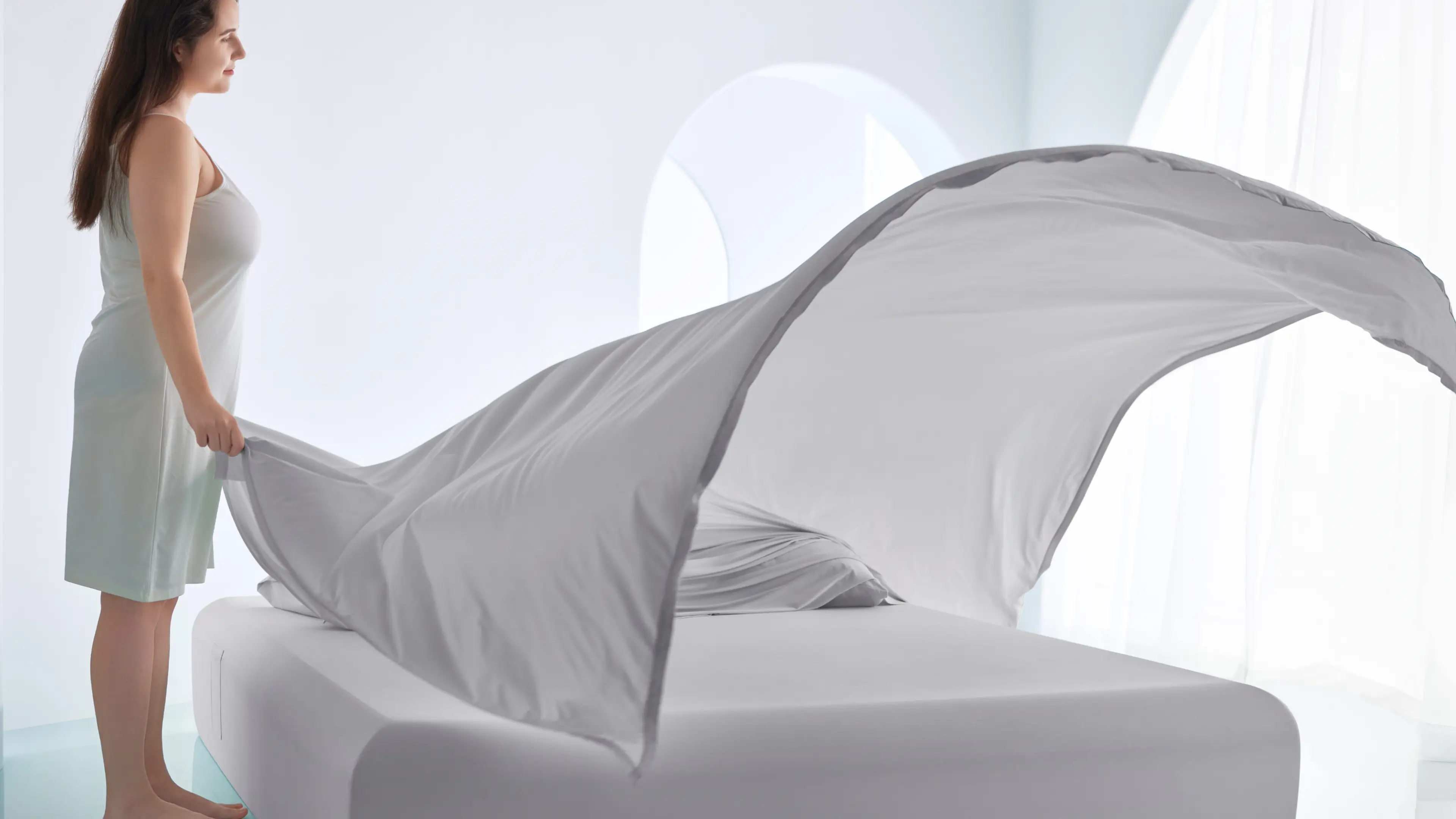 A woman in a nightgown spreads a large, billowing white sheet over a bed in a bright, airy bedroom