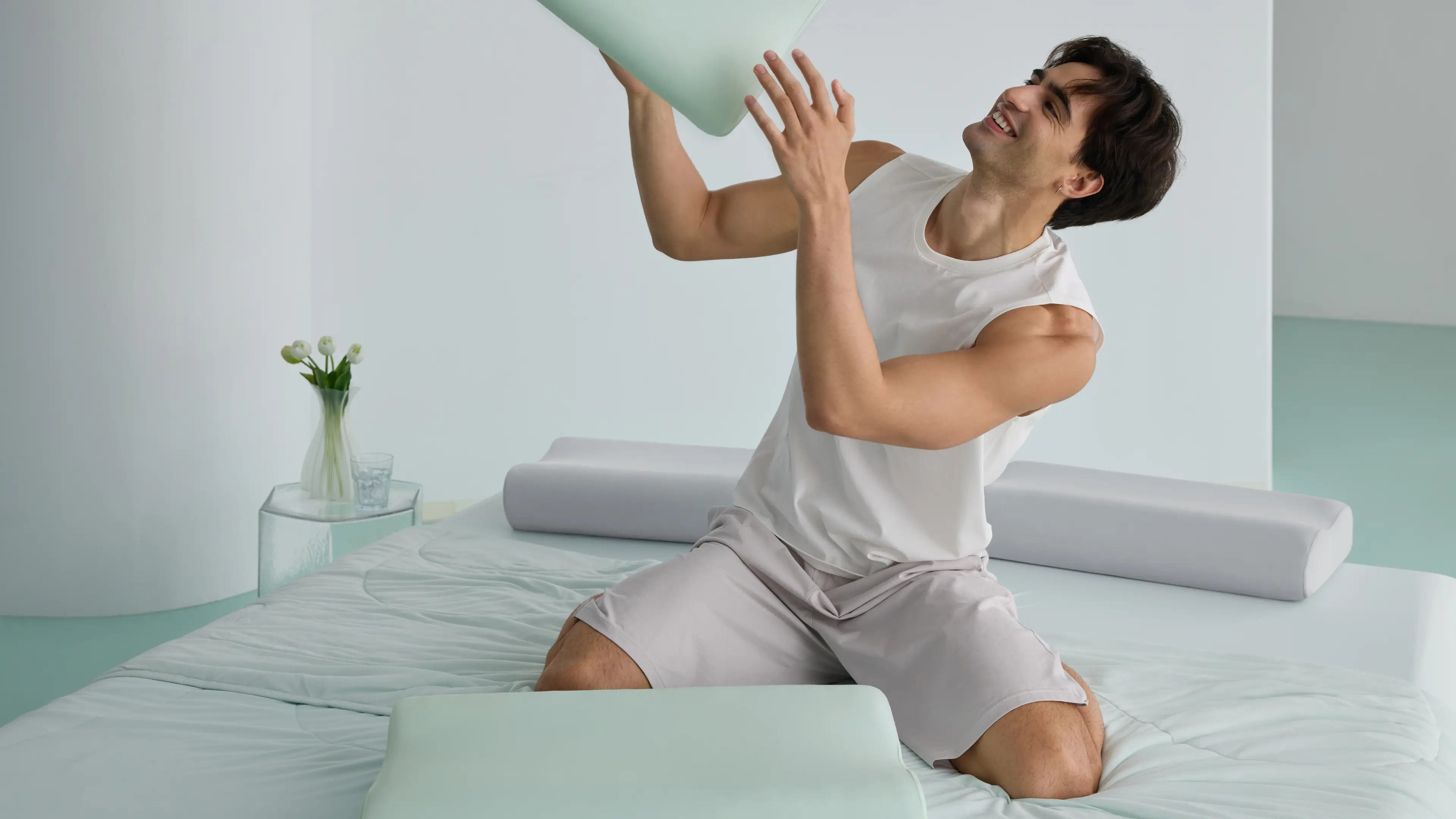 Smiling man kneeling on a bed, playfully lifting a light green memory foam pillow in a modern white bedroom