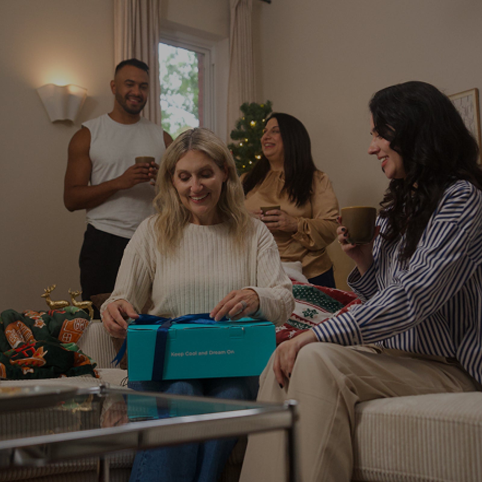 Two women opening a blue box together in a living room with two other people in the background.