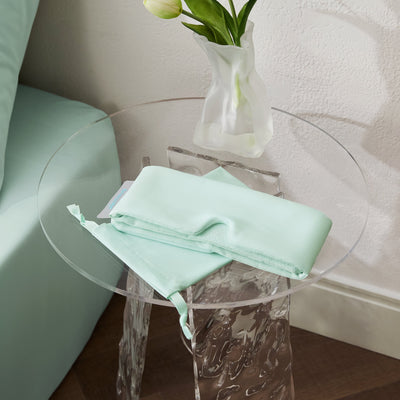 Mint green Breescape cooling eye mask folded on a clear table with a vase of flowers.